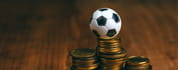 La imagen muestra pilas de monedas con un balón de fútbol pequeño coronándolas.